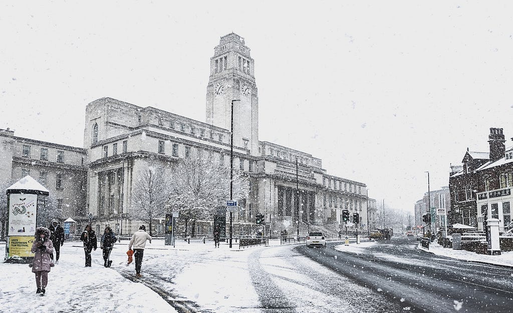 The Parkinson Building including the large clock tower, viewed from Woodhouse Lane. The paths and buildings are lightly covered in a recent snow shower. People are walking up the street.