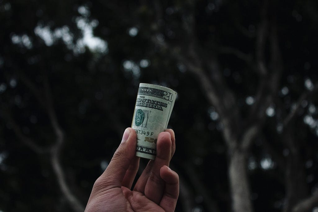 A hand holding a roll of money with lush, green trees in the background