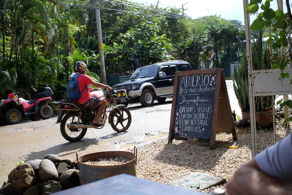 The view from a coffee shop with a motorcyclist driving by on a dirt road.