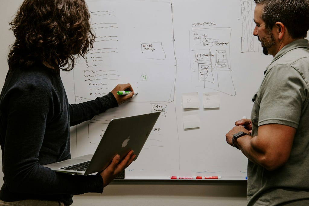 Two designers engaged in a collaborative session at a whiteboard, where one is holding a laptop and the other is holding a marker, indicating an interactive design planning process.