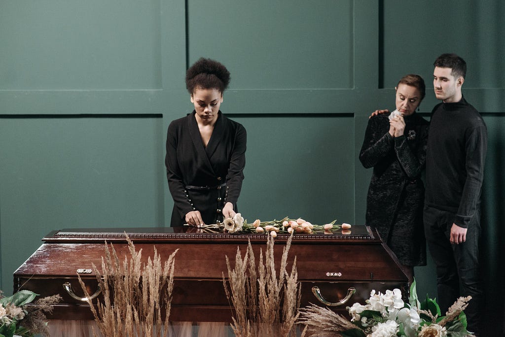 Female person of color standing over a casket while a white male comforts a white female in the corner.