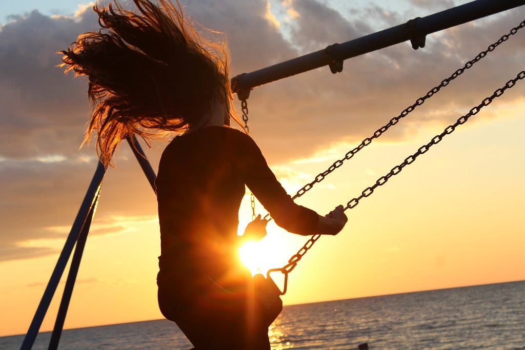 Photo of a person on a swing, their long hair trailing behind them as they swing. The background is a sunset along above the water.