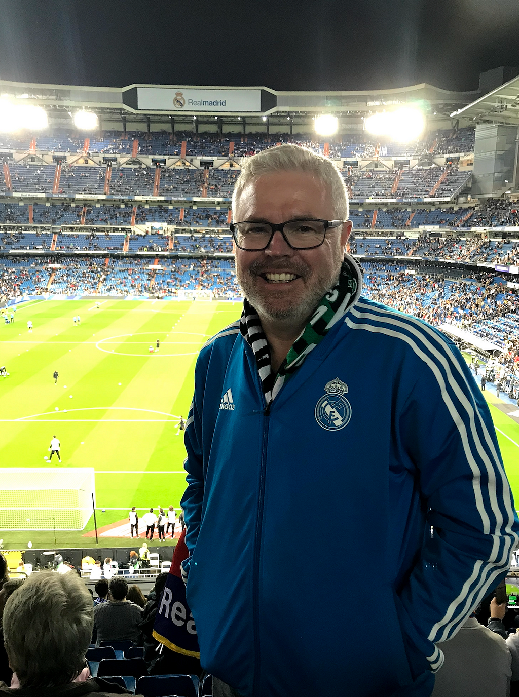A man in a Real Madrid jacket smiles in front of the soccer field.