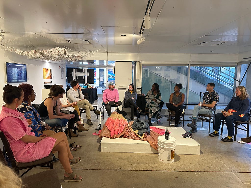 A group of people gather in chairs in a roundtable format in a white room with floor to ceiling windows. They wear bright pinks and dark colors. In the center of the table and overhead are tapestry-based sculptures.