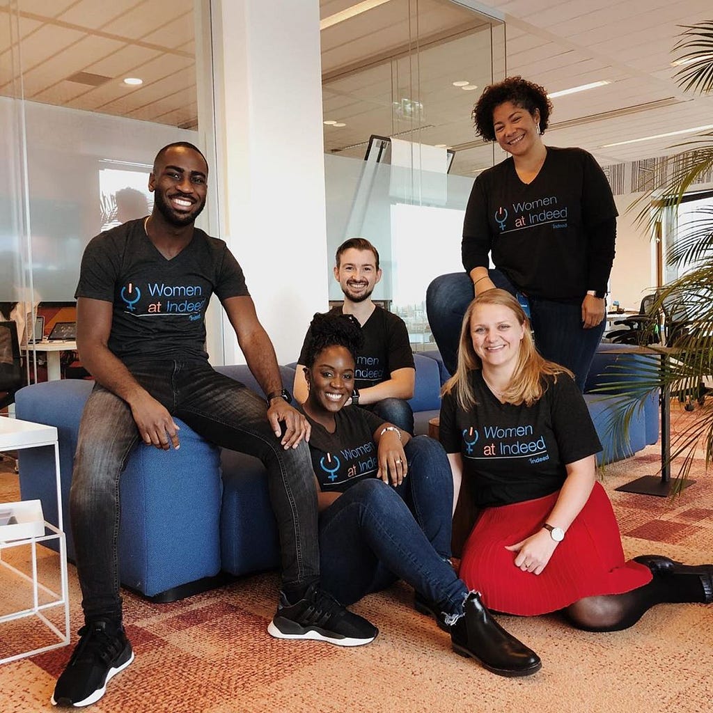 Image of five Indeed inclusion group members smiling and wearing shirts labeled “Women at Indeed” in a relaxed office setting