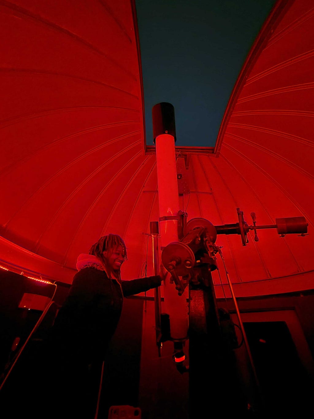 Black girl smiling next to telescope in observatory illuminated with red light. The telescope is in a window where the sky is cloudy