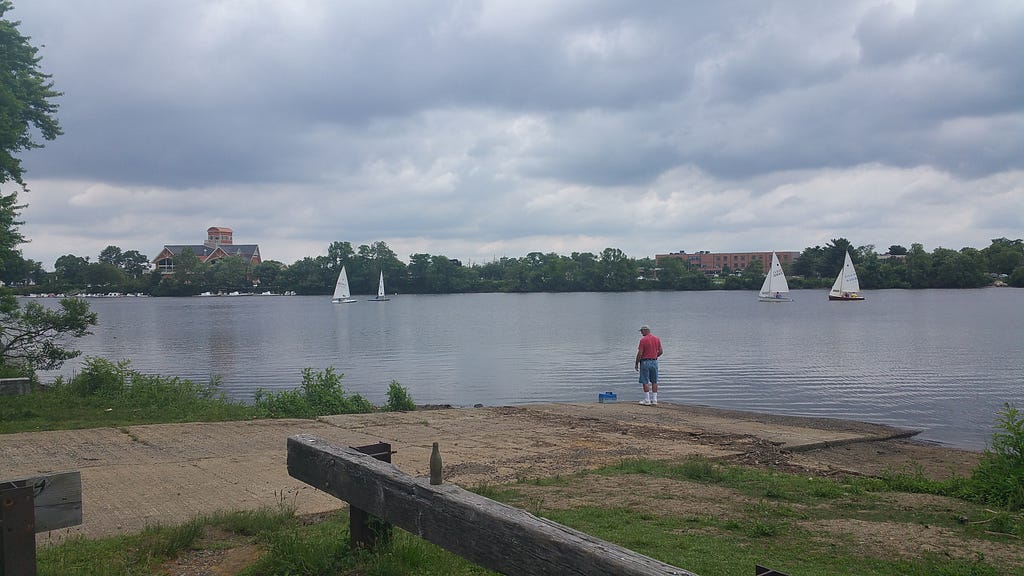 a man fishes from a boat ramp along the river as sail boats pass
