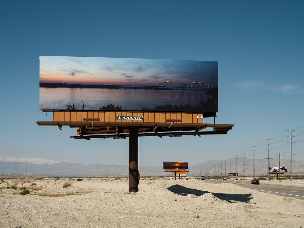 The sandy shoulder of a highway with rows of billboards on either side. In the foreground is a billboard covered by a sunset photo of a body of water, with a bridge off in the distance.