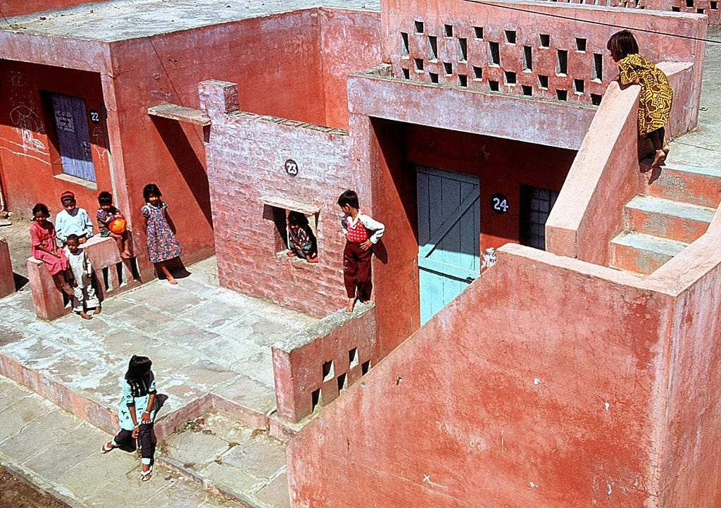 Under bright sun light, kids gather in the front yard of the single story building made of bright red clay