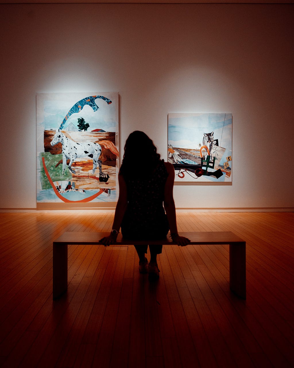 A woman is sitting alone in an art gallery, looking at two paintings on a wall.