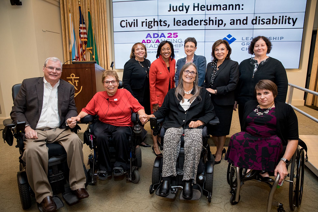 9 people—5 in back, 4 in front—smile for the camera. They are professionally dressed. The 4 in front are using wheelchairs.