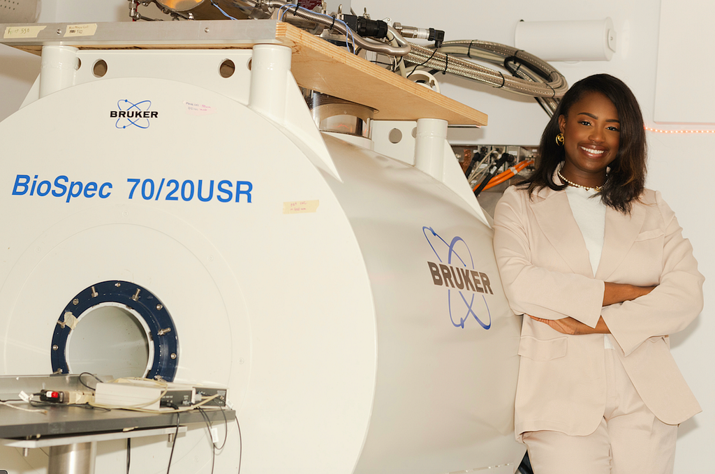 Black woman next to a big MRI machine that says BioSpec 70/20USR and a Brucker logo. The woman is wearing a tan blazer and has gold accessories. She is smiling ear-to-ear and has her arms folded in front of her.