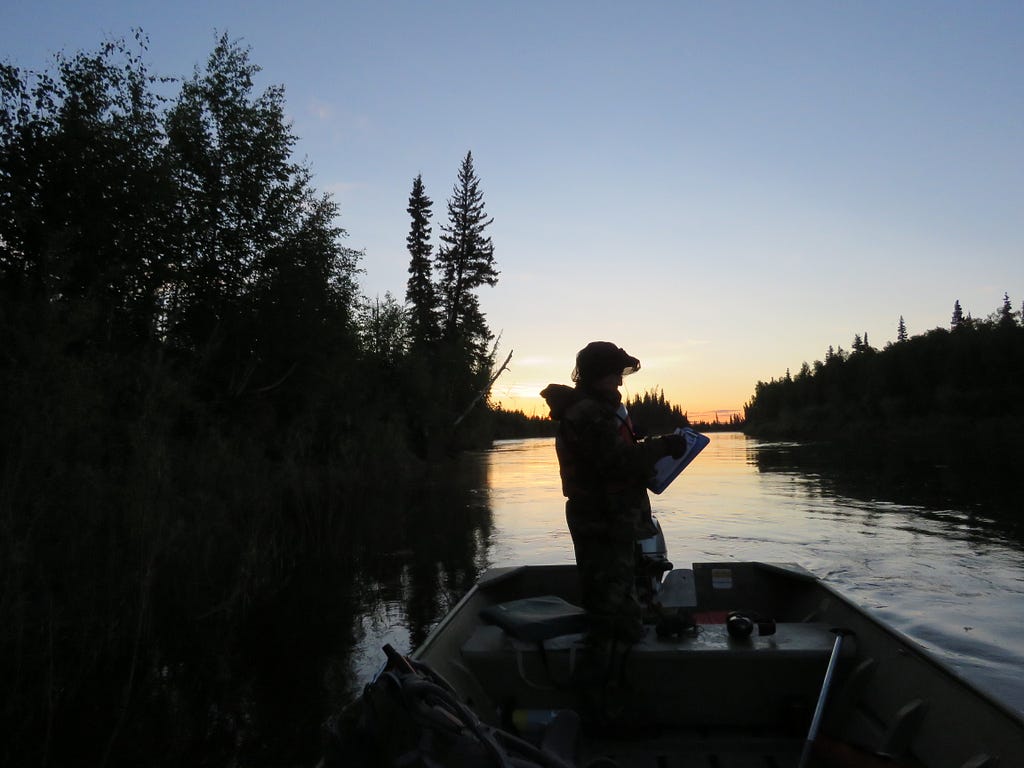 A person stands in a boat in silhouette with forest and water and the beginning of a sunrise in the background.