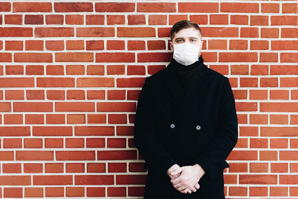 A man in a surgical mask stands against a brick wall.