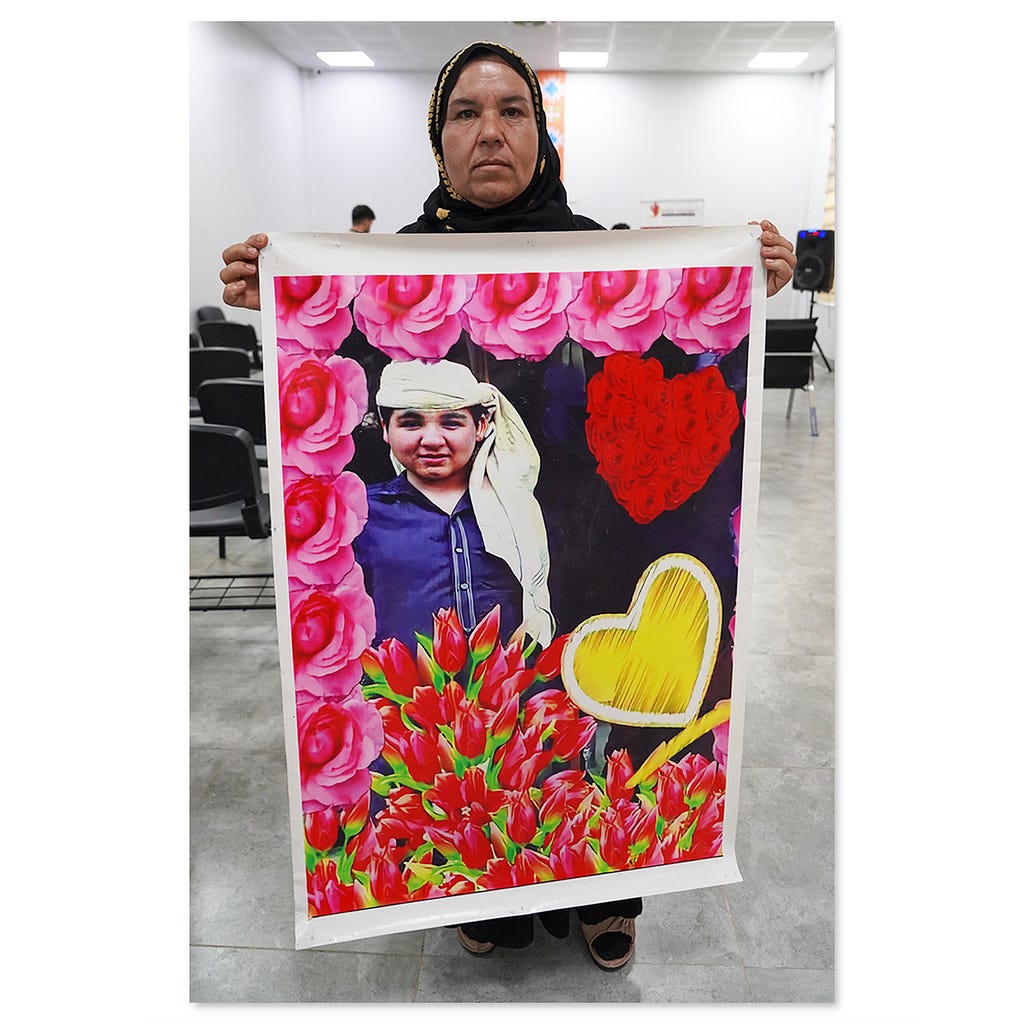 A woman wearing a head scarf holds a large photograph of a boy surrounded by flowers.