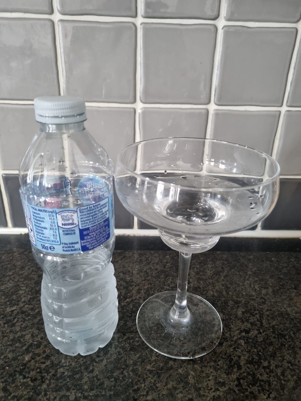 Bottle of water on a table with a cocktail galss with water next to it. Dink more water — flushes toxins from the body.