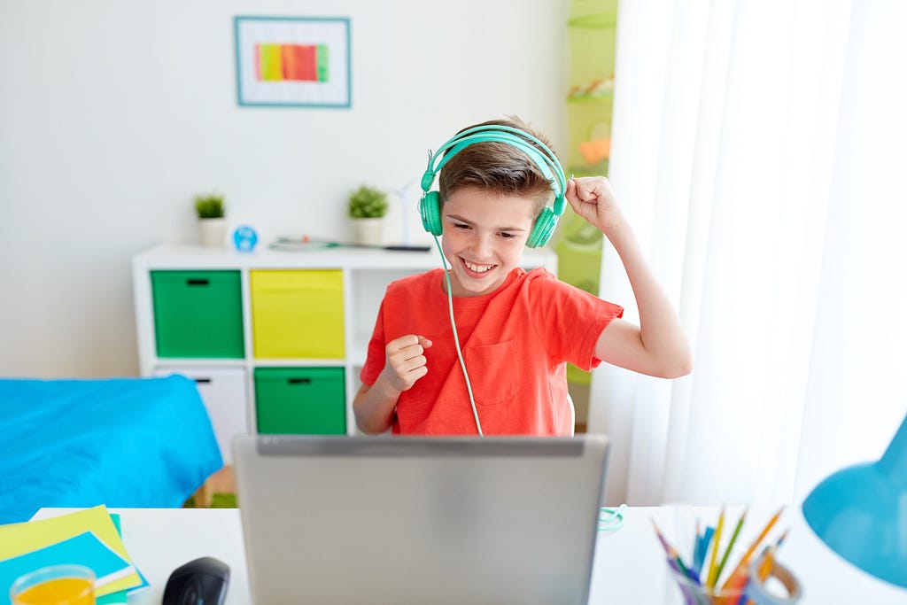 A boy in red T-shirt and green headphones, expressing the feeling of victory after winning a video game on a laptop.