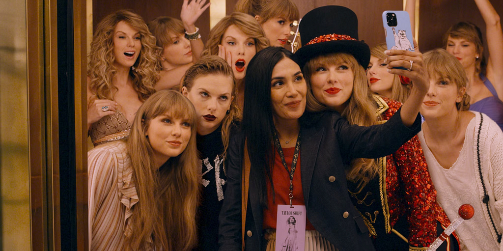 A woman in the elevator taking a selfie with a bunch of Taylor Swifts in different outfits.