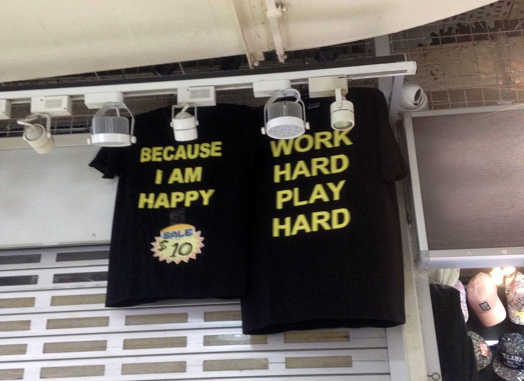 Two black shirts on sale that are hung at some mall with the writings ‘Because I am happy’ and ‘Work Hard Play Hard’ in each shirt.