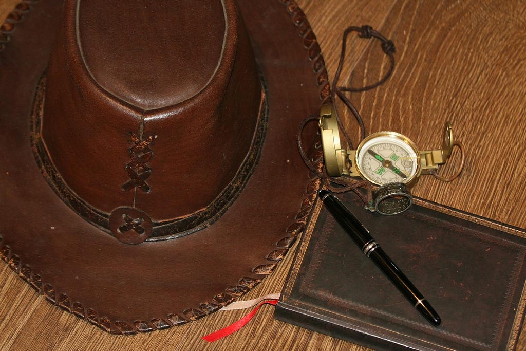 Objects on a table suggesting exploration and adventure: a hat, a compass, a notebook and a pen.