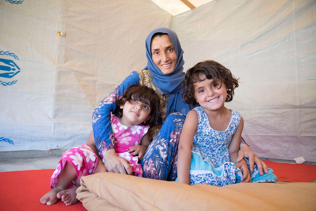 A woman dressed in blue holds her two children, age 4 and age 3, close. They are all smiling. They are seated on the floor inside what appears to be a tent or makeshift shelter.