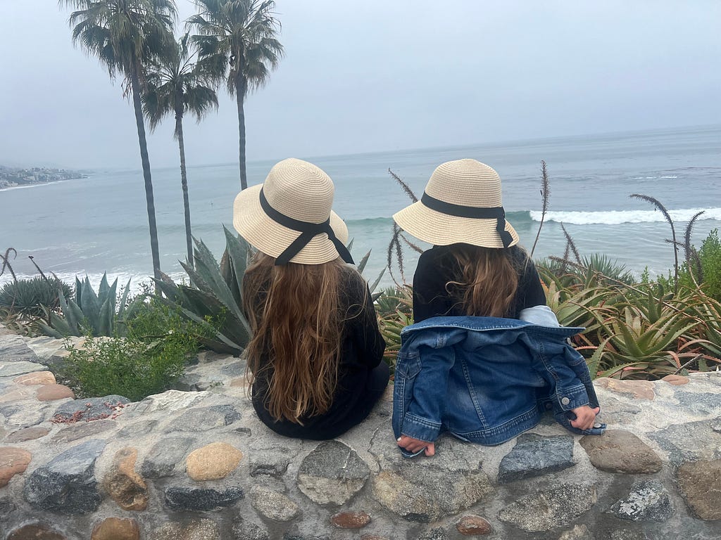 Two young girls sit on a stone wall overlooking the Pacific ocean.
