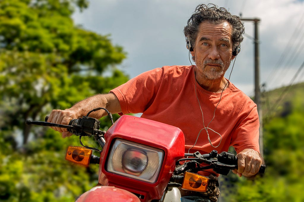 Ator Chico Díaz dirige uma moto vermelha. Ele usa fones de ouvido e uma camiseta laranja. Ao fundo, há árvores e um poste com fios de energia.