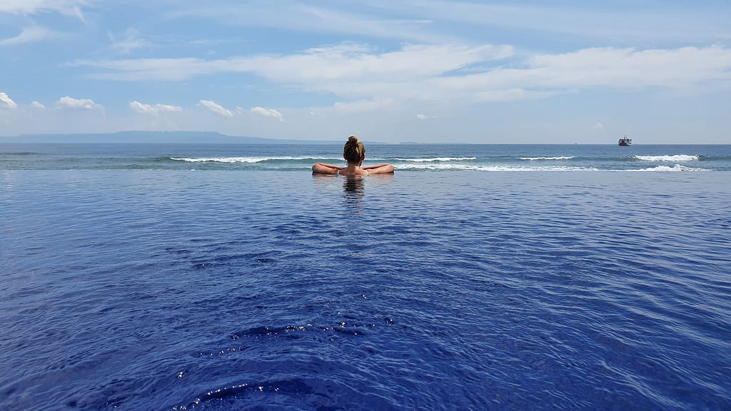 The view from an infinity pool, of a woman overlooking a Balinese beach.