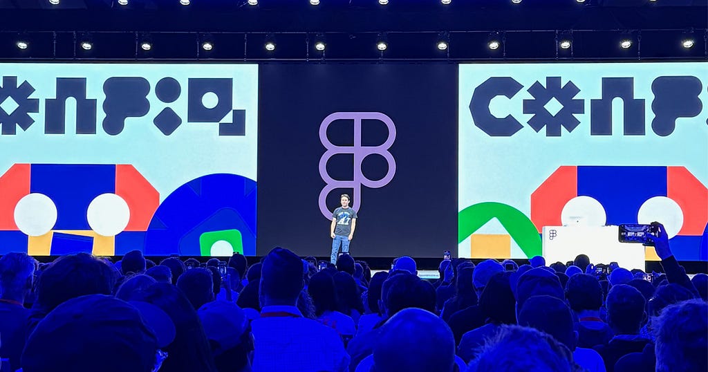 Figma CEO Dylan Field speaking on stage at Config, a large conference with an audience seated facing the stage. The stage backdrop features colorful geometric designs with the Config logo prominently displayed in the center. The speaker is standing in front of a large audience seated in rows facing the stage. The stage is well-lit with spotlights, creating a professional and engaging atmosphere for the conference presentation.