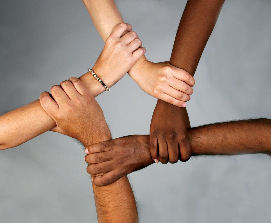 Five hands of people from varying races holding each other’s wrists forming a pentagon shape in the middle.