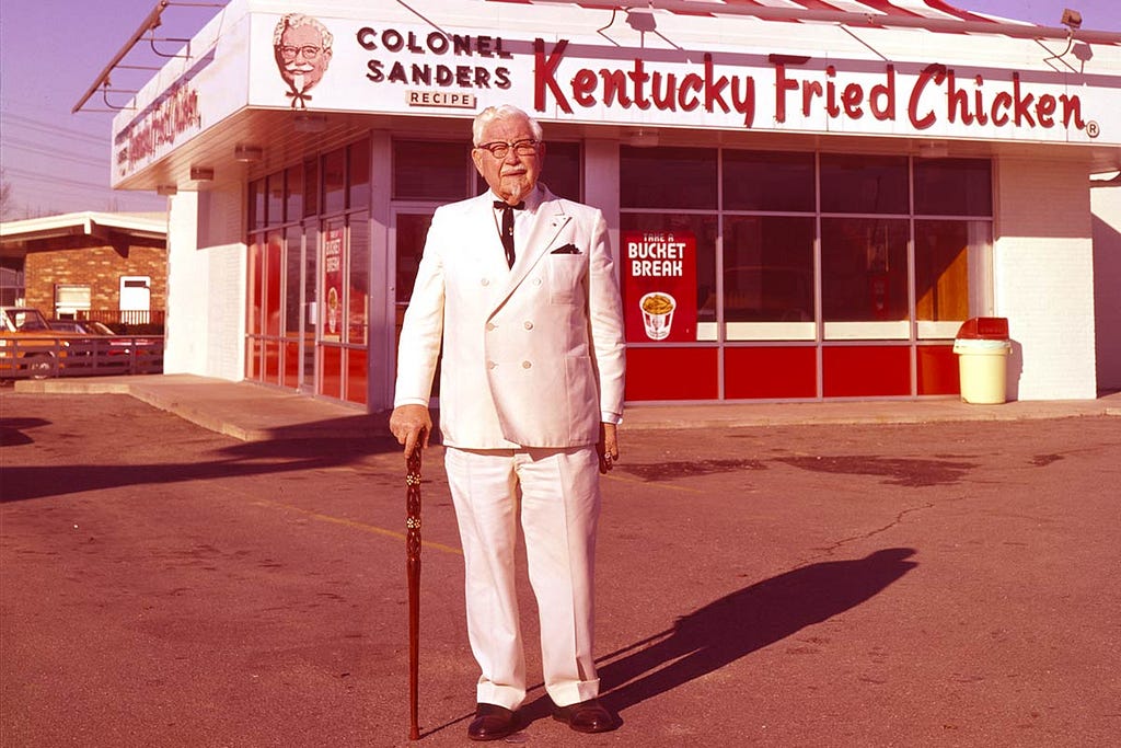 A photo portrait of Harland Sanders, in front of his restaurant “Kentucky Fried Chicken.