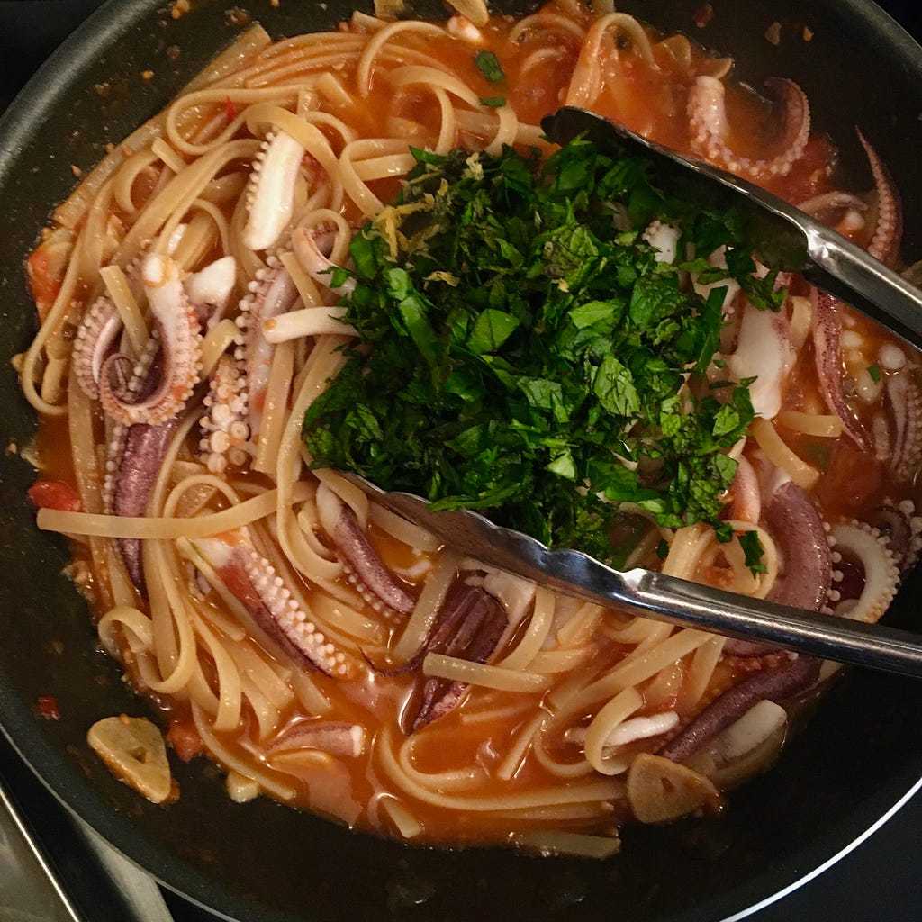 Overhead shot of a pan of linguini in sauce with squid tentacles on top, being topped with chopped herbs.