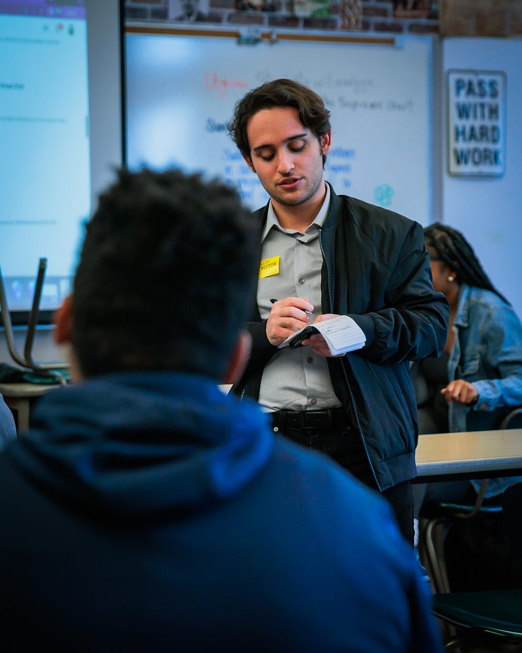 A reporter writes in a notepad while he interviews a high school student in a classroom.