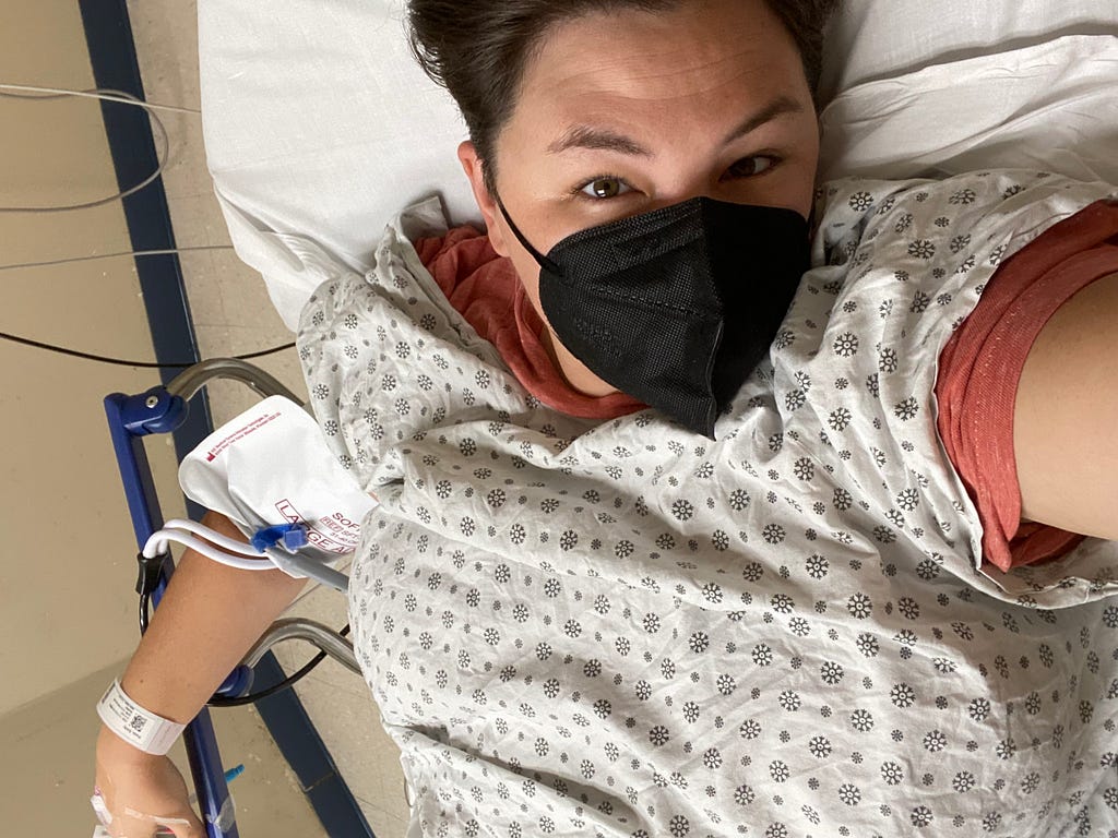 A white woman in her thirties takes a selfie of herself in a hospital bed. She is wearing a hospital gown over her shirt and a mask on her face, and she is hooked up to lots of tubes