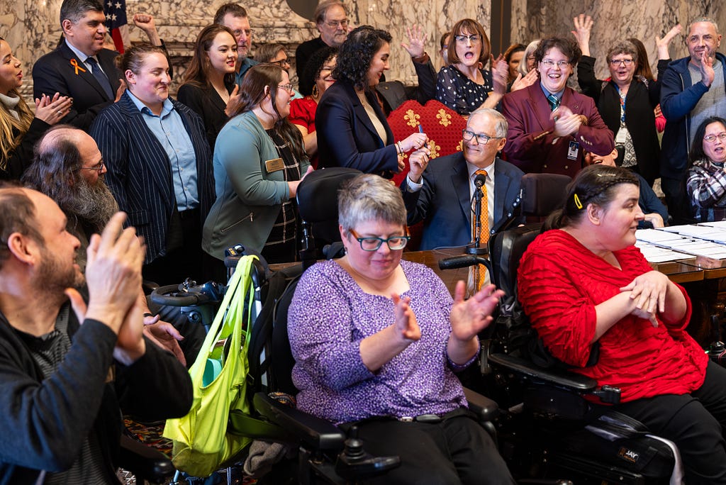 The image shows a bill signing. A group of people surround the governor and the large table at which he sits. The governor is wearing a blue suit with an orange tie and he is sitting in a chair upholstered in crimson and gold fabric. He is handing a pen to the woman on his right. The crowd of people (some of whom are in wheelchairs) are clapping and celebrating.