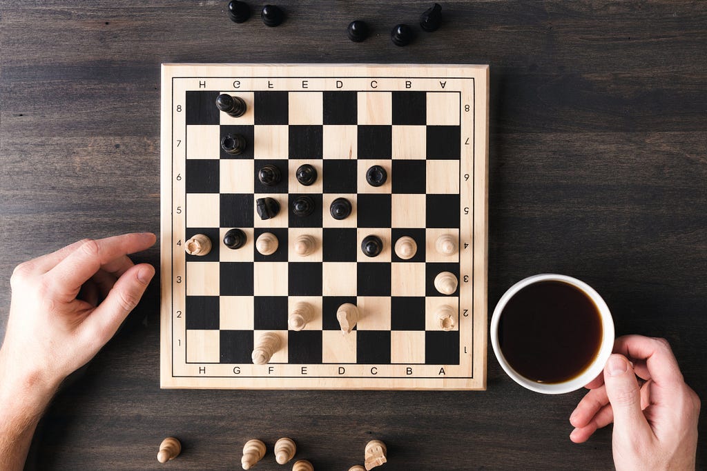 A chess game on a small board framed by two hands and a cup of coffee.