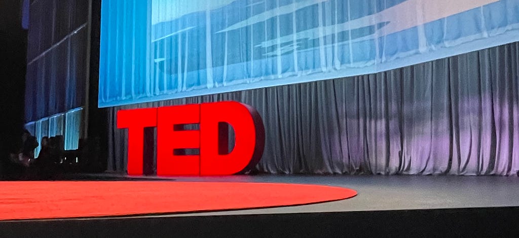 A photograph of the TED stage from the front row of the audience, showcasing the red dot carpet and the big red TED letters in front of a curtain.