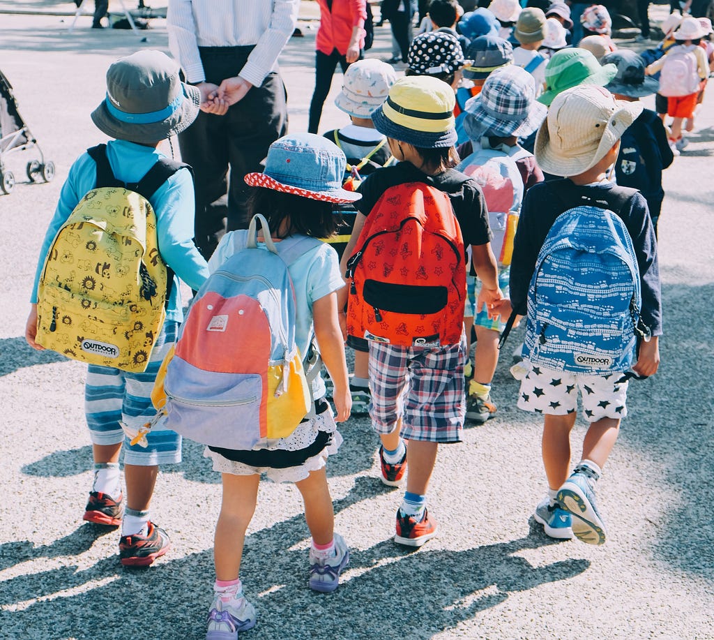 Young school children crossing a street from the back, multi-color backpacks and wide brimmed hats can be seen.