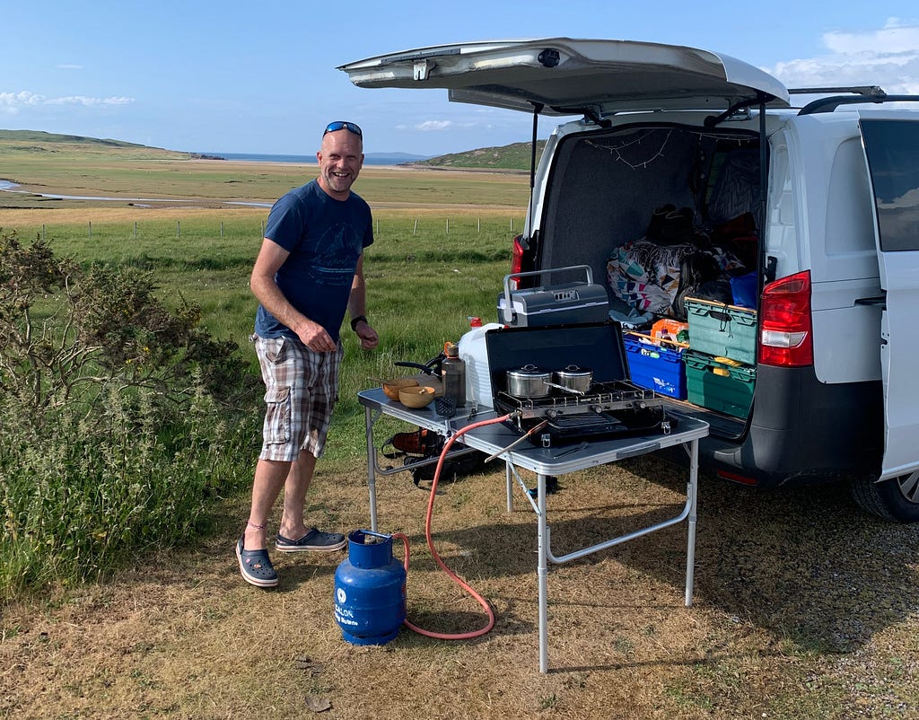 Duncan stands beside his camping cookset outside his van loaded with camping supplies.