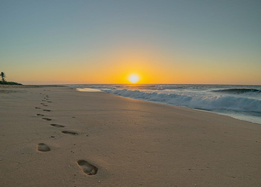 A landscape featuring beach and surf and a perfectly clear sky. A line of footprints in the sand stretches backwards from the viewer along the beach towards the setting sun