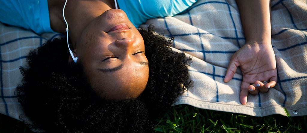 A woman wears headphones and lays down on a blanket in a park, smiling serenely.