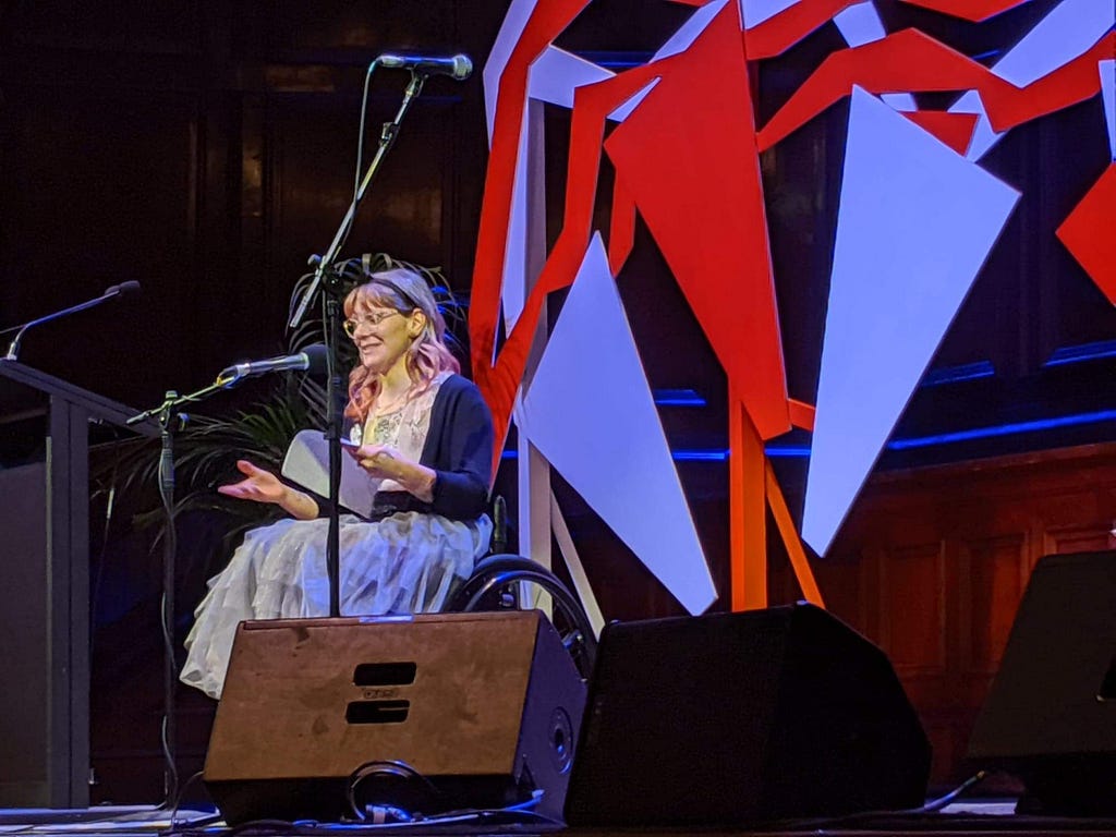 image of a woman in a wheelchair on stage reading, she is wearing a tutu, glasses and has long pink curly hair