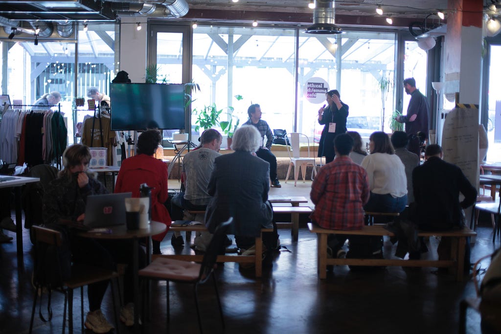 A speaker is standing and holding a microphone, whilst talking to another who is seated. They are in front of a large window that has a small round sticker that reads “Ethical Dilemma Cafe”. Clusters of audience members are seated on chairs and wooden benches in front of them.