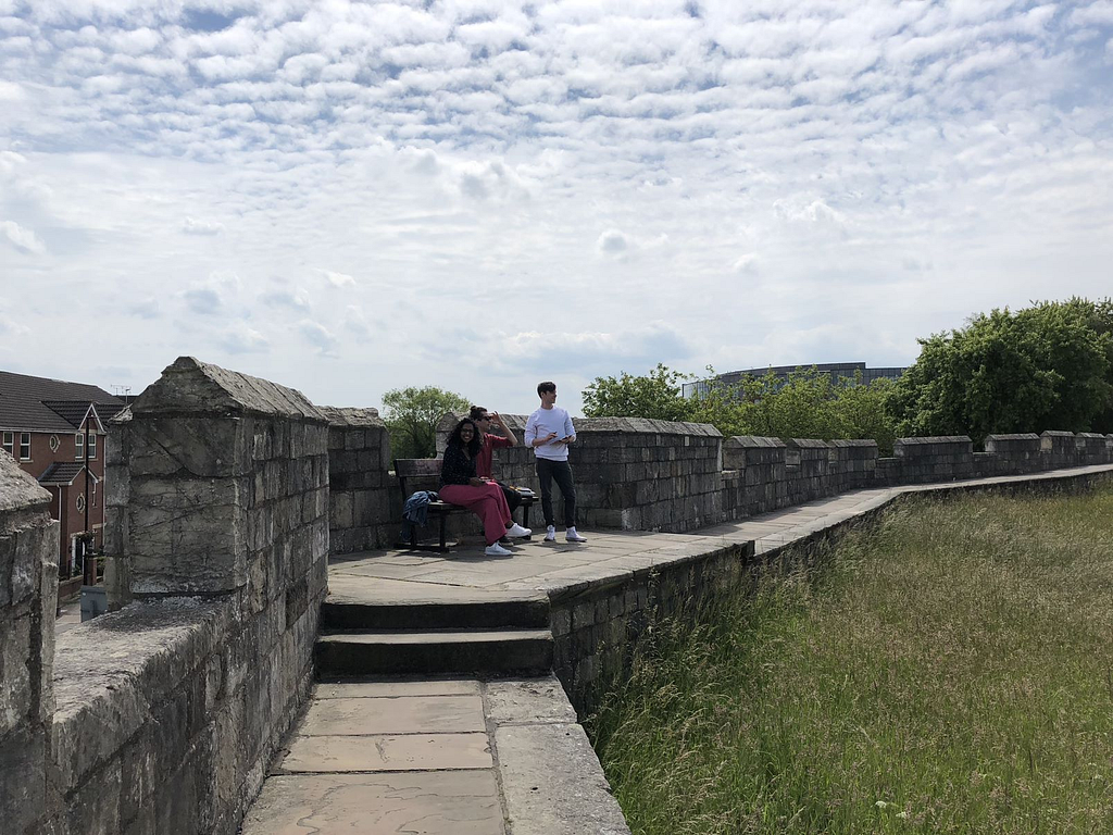 In the middle of this picture are three people: two sitting on a bench and one standing up. They are on the York Wall, a medieval stone fortification. You can see some clouds and bright blue sky above them.