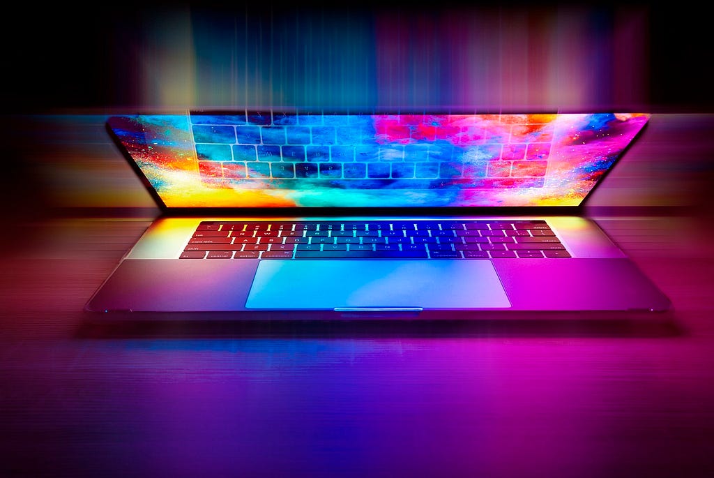 A laptop sits partially opened on a semi-reflective surface. The laptop monitor displays a bright multi-colored graphic and also displays a reflection of the keyboard. The image has been edited to bleed the different colors from the monitor in all directions to the edge of the image.