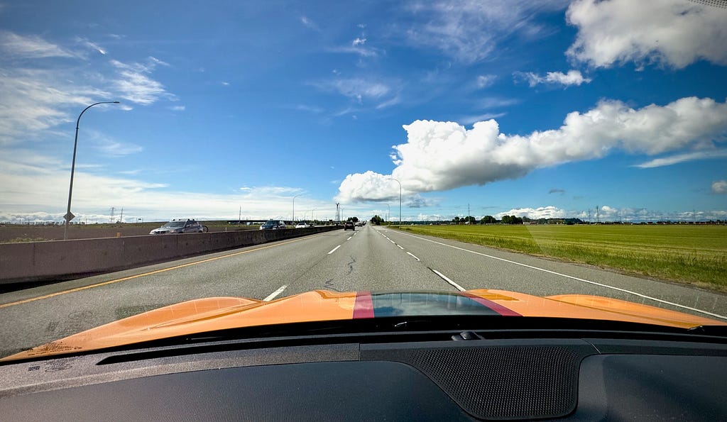 Looking down the 3-lane highway stretching out under blue skies and fluffy clouds with a green field on the right