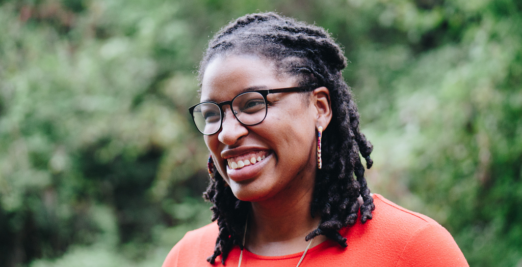 A black woman with glasses smiling, there’s lots of greenery in the backgound