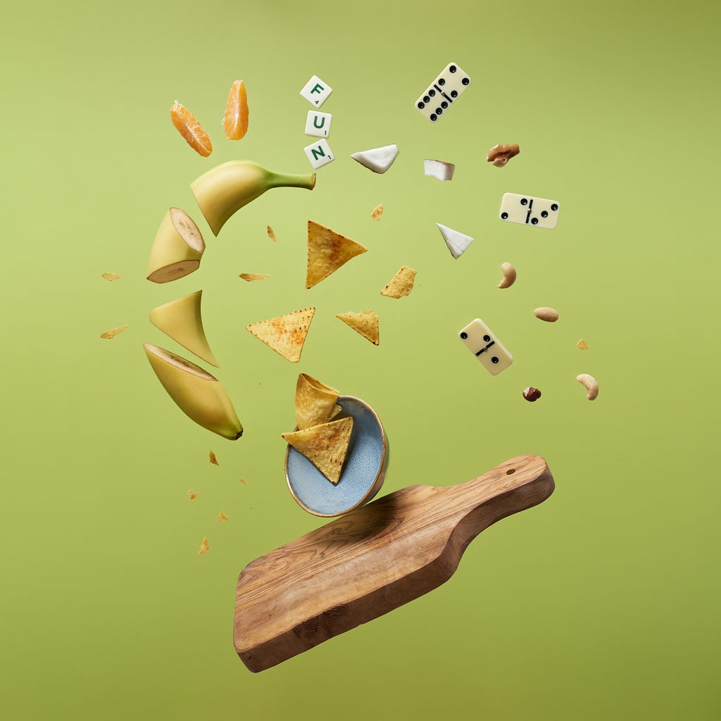 A banana chopped in random pieces tossed up in the air with dominoes, nachos and some scrabble alphabets along with it from a wooden chopping board.