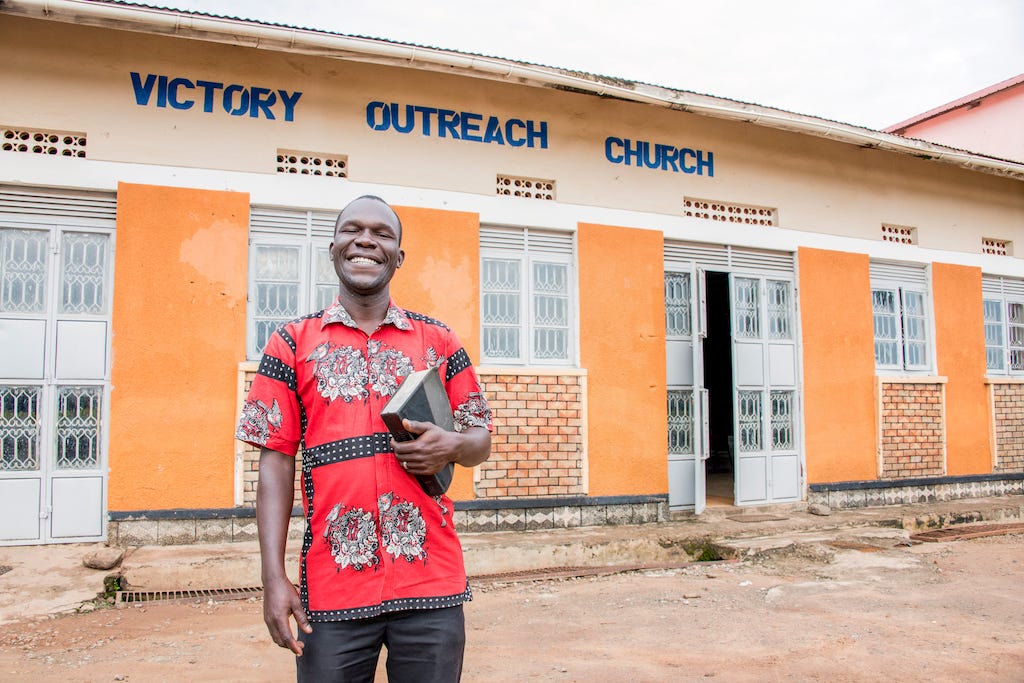 A man in a red patterned shirt stands smiling and holding a Bible in front of Victory Outreach Church, a yellow building.