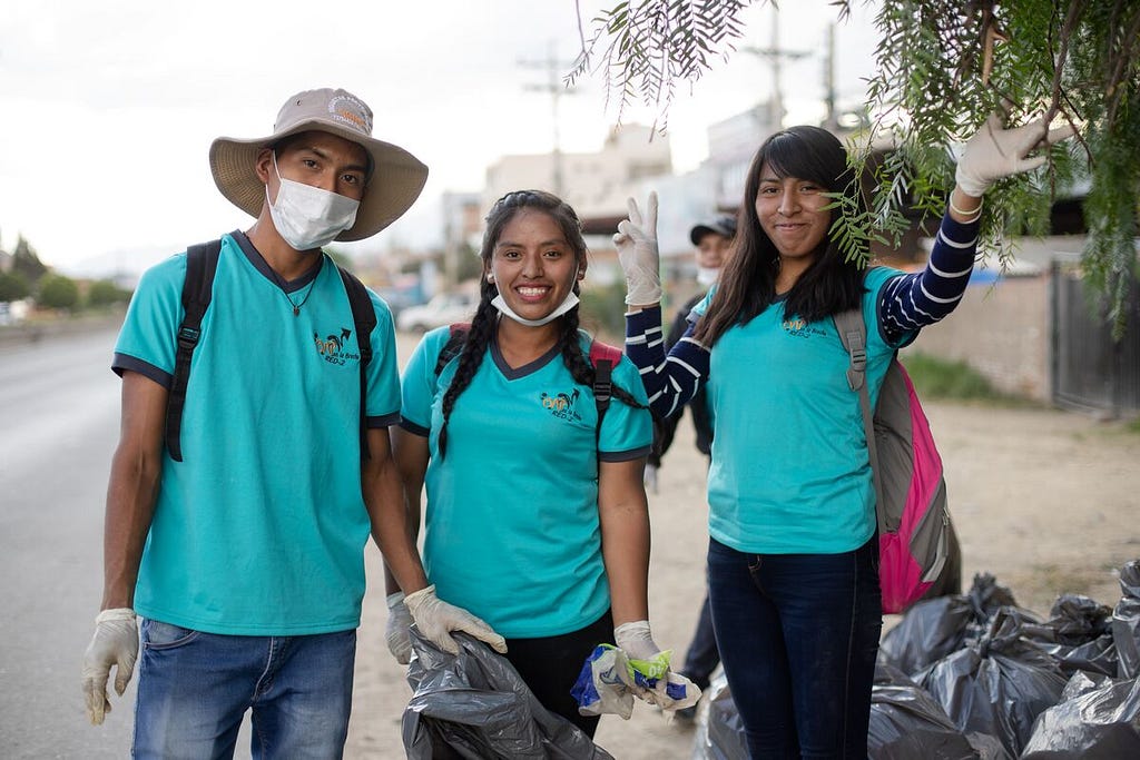 In Bolivia, three teens, two girls and a boy, are wearing blue t-shirts and are taking a break from picking up trash.
